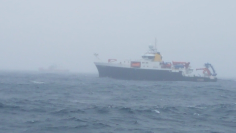 This image shows one of the research ships floating in rough seas in the foreground with another ship barely visible in the background on the left side of the frame. The weather is rainy with a very gray sky. The top half of the ship is white and the lower half is black. Orange or blue cranes are visible on bow and stern of the ships. 