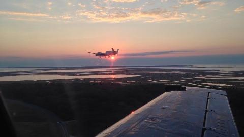 This image shows a Delta Hawk drone as seen from the window of a chase plane. The drone is positioned in the middle of the frame and flying over a marine marsh area at sunset. The Sun is just below the bottom of the drone and about to disappear behind a cloudbank on the horizon. The right wing of the chase plane can be seen extending from the lower-right corner of the image.