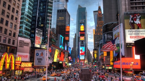 This image features a view of New York City's Times Square looking south in early evening. The right, left, and middle background of the image shows office buildings, skyscapers, and stores with bright lights and advertising signs surrounding the square. The center of the image shows cars and people traversing the streets and sidewalks below.