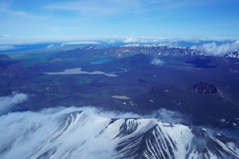 This is an aerial image of Okmok caldera. Inside the caldera are cinder cones and lakes. The rim of the caldera is covered with snow. The caldera is ringed by the surrounding island and ocean. Over the crater is blue sky with thin clouds.