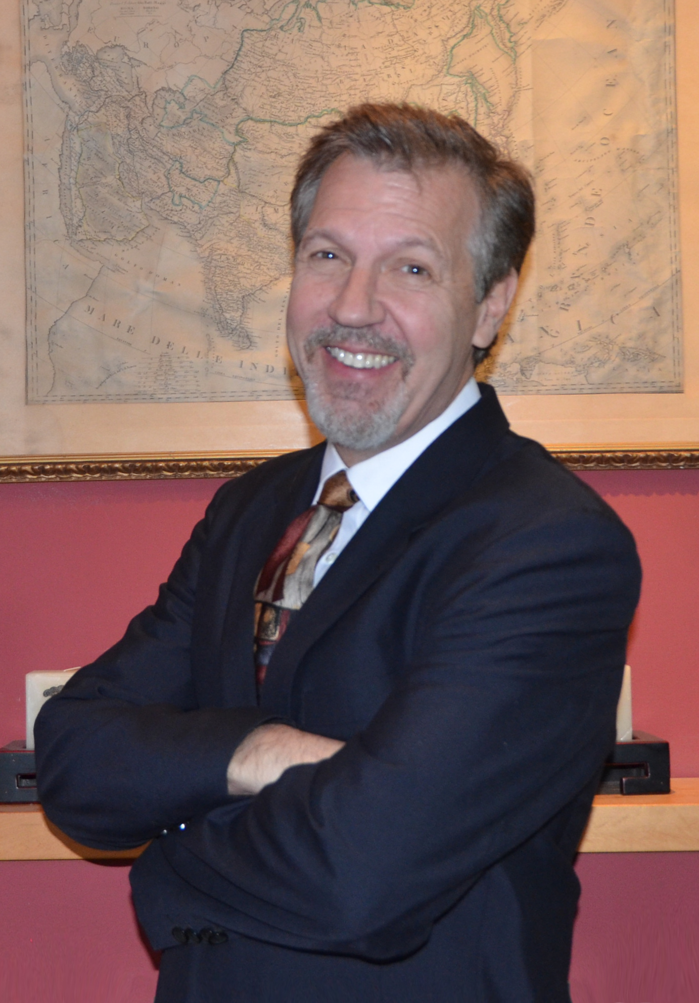 Headshot of Drew Kittel wearing a blue suit and striped necktie and standing in front of an old map of china.